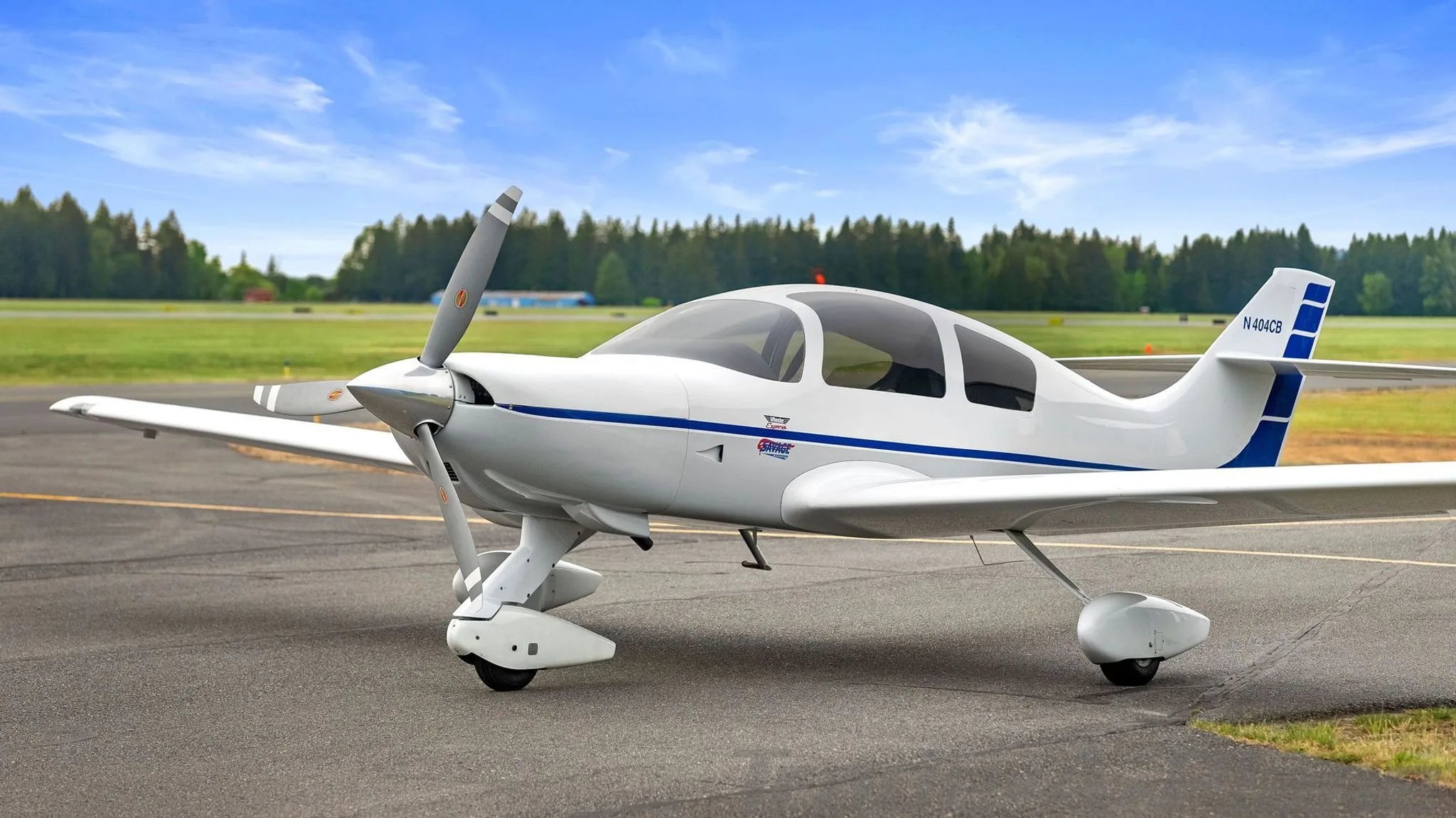 A white single-engine aircraft with blue stripes on the fuselage and tail, registration N404CB, parked on an asphalt runway with trees in the background.