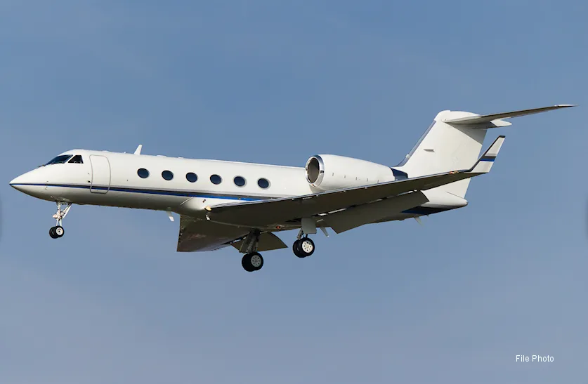 A white Gulfstream business jet with blue accents, landing gear extended, flying against a clear blue sky.
