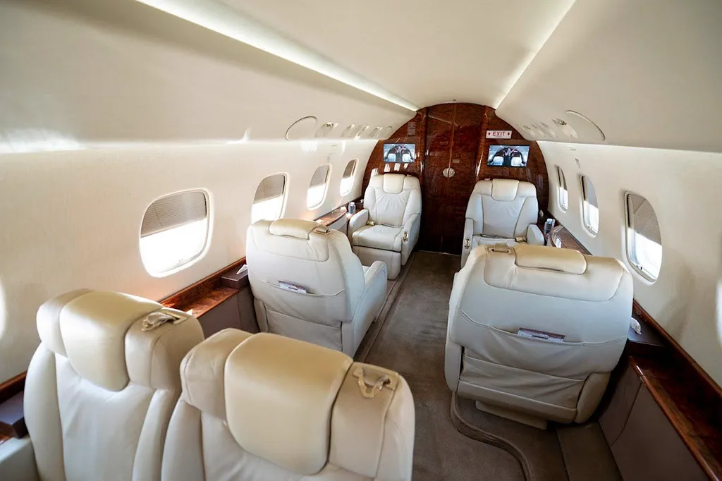 The forward section of a business jet cabin with beige leather seats and two monitors on the bulkhead.