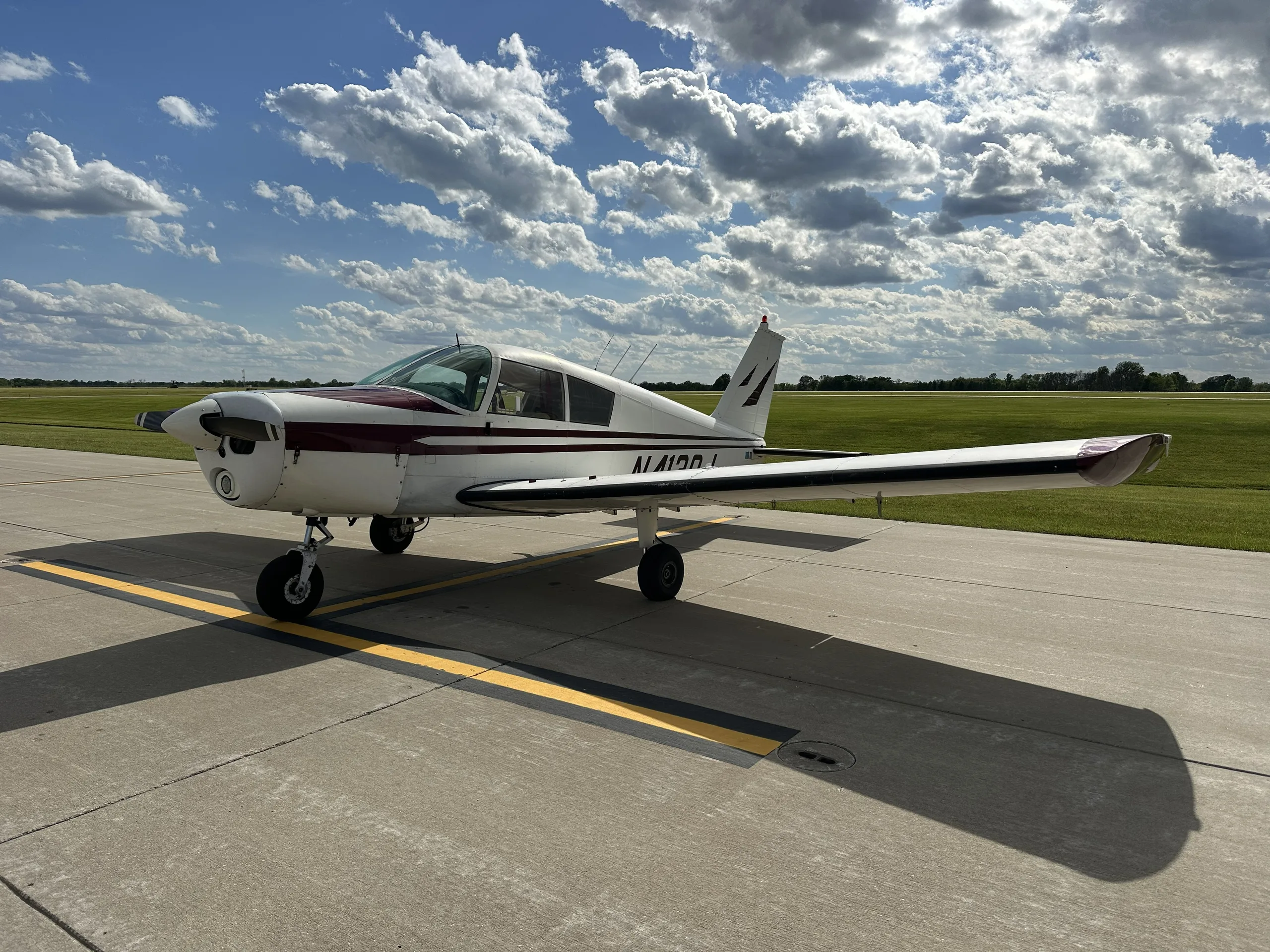 A white and maroon single-engine aircraft, likely a Piper Cherokee, parked on an asphalt ramp with green grass in the background under a partly cloudy sky.