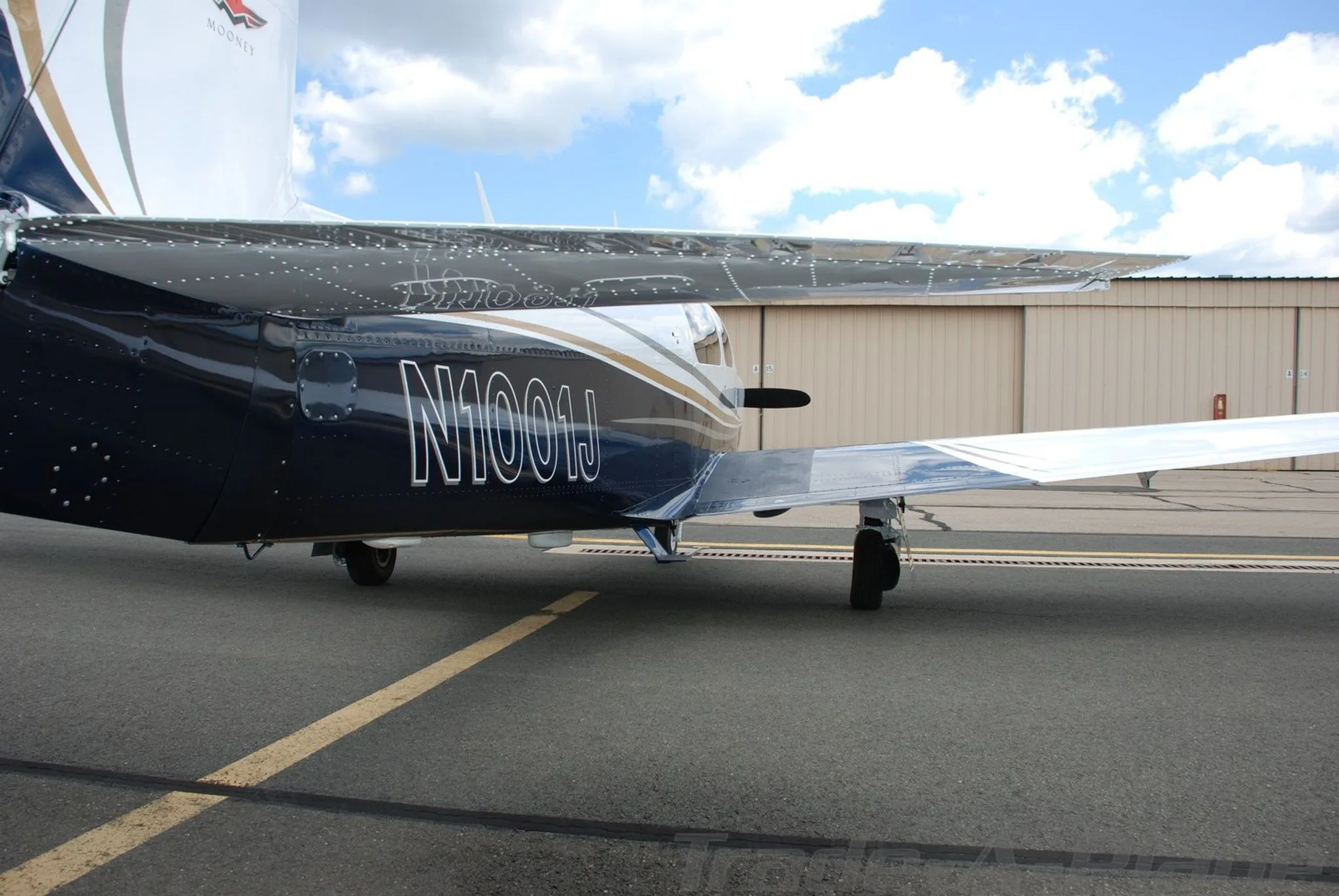 A close-up view of the fuselage and wing of the white and blue Mooney aircraft (N1001J) on a tarmac.