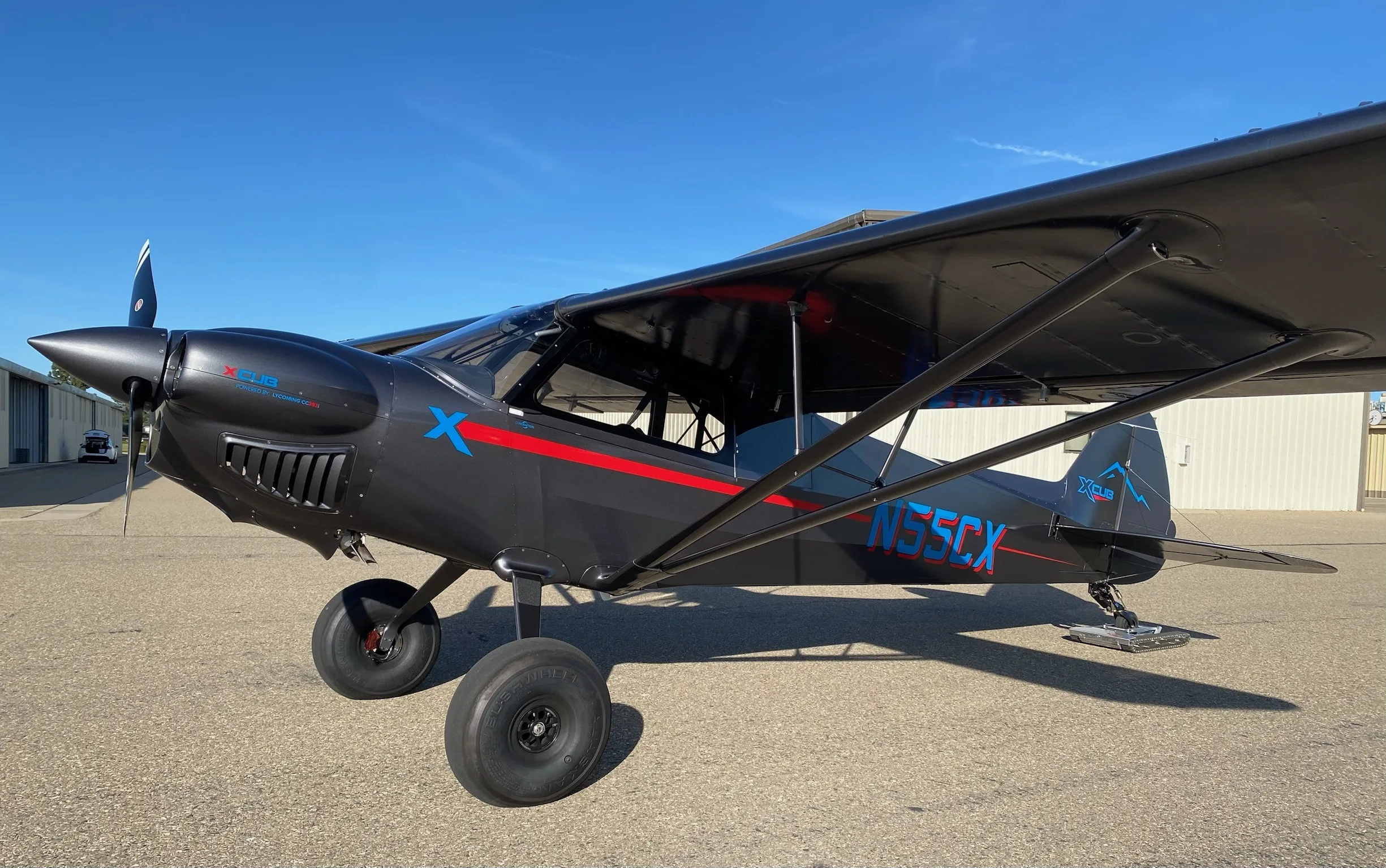 A dark gray CubCrafters X-Cub with red and blue accents, large bush tires, and a tail ski is parked on an asphalt tarmac.