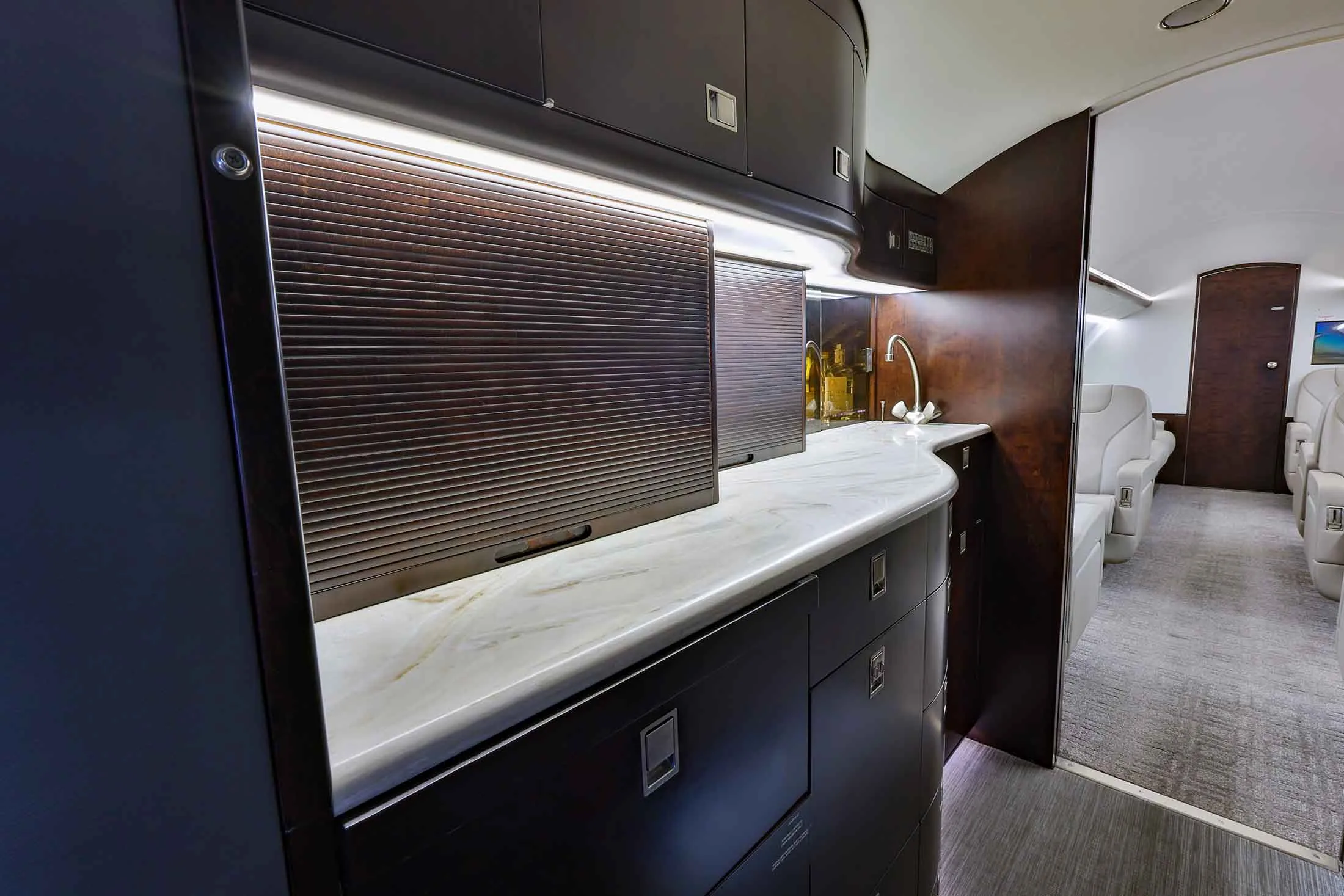 A luxurious aircraft galley with dark wood cabinetry, a white marble countertop, and a sink, leading to the passenger cabin.
