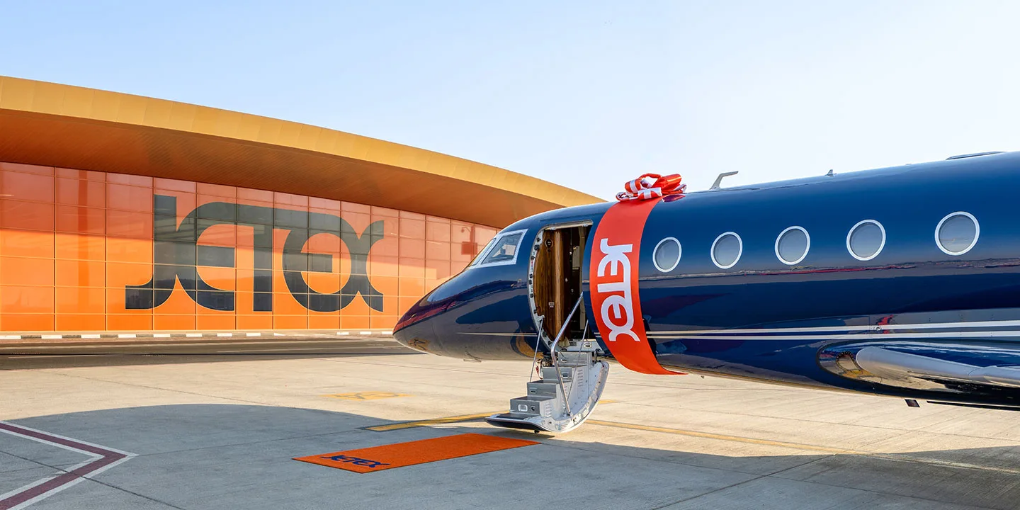 A dark blue business jet with an orange 'Jetex' ribbon and steps down, parked in front of a large orange building with 'Jetex' branding.