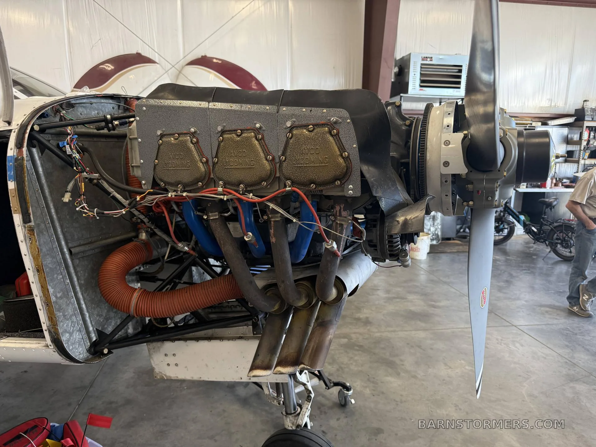 The engine bay of a Piper Cherokee Lance (N75398) with the cowling removed, showing the engine, exhaust, and propeller in a hangar.