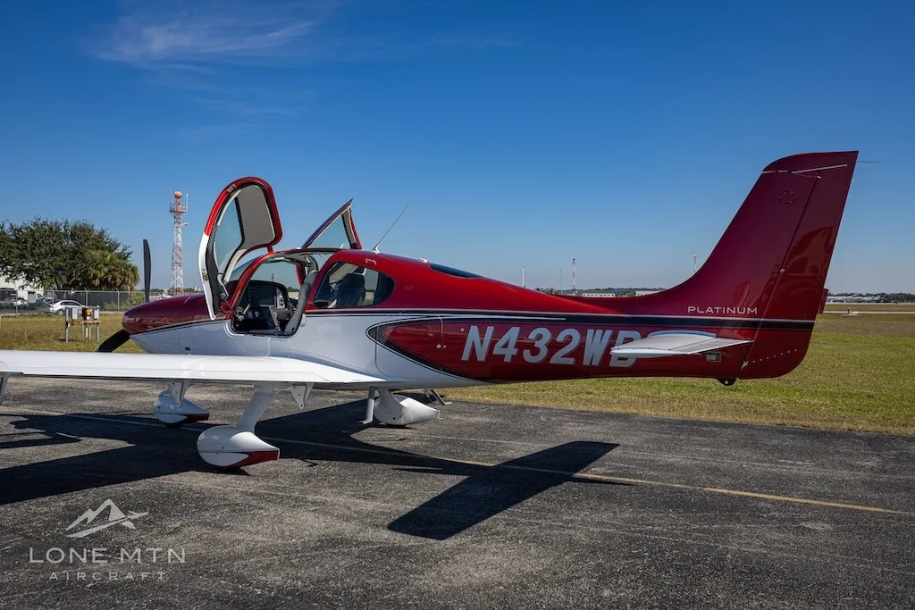 A side view of the red and white Cirrus SR20 aircraft (N432WB) with its doors open, parked on an airport tarmac.