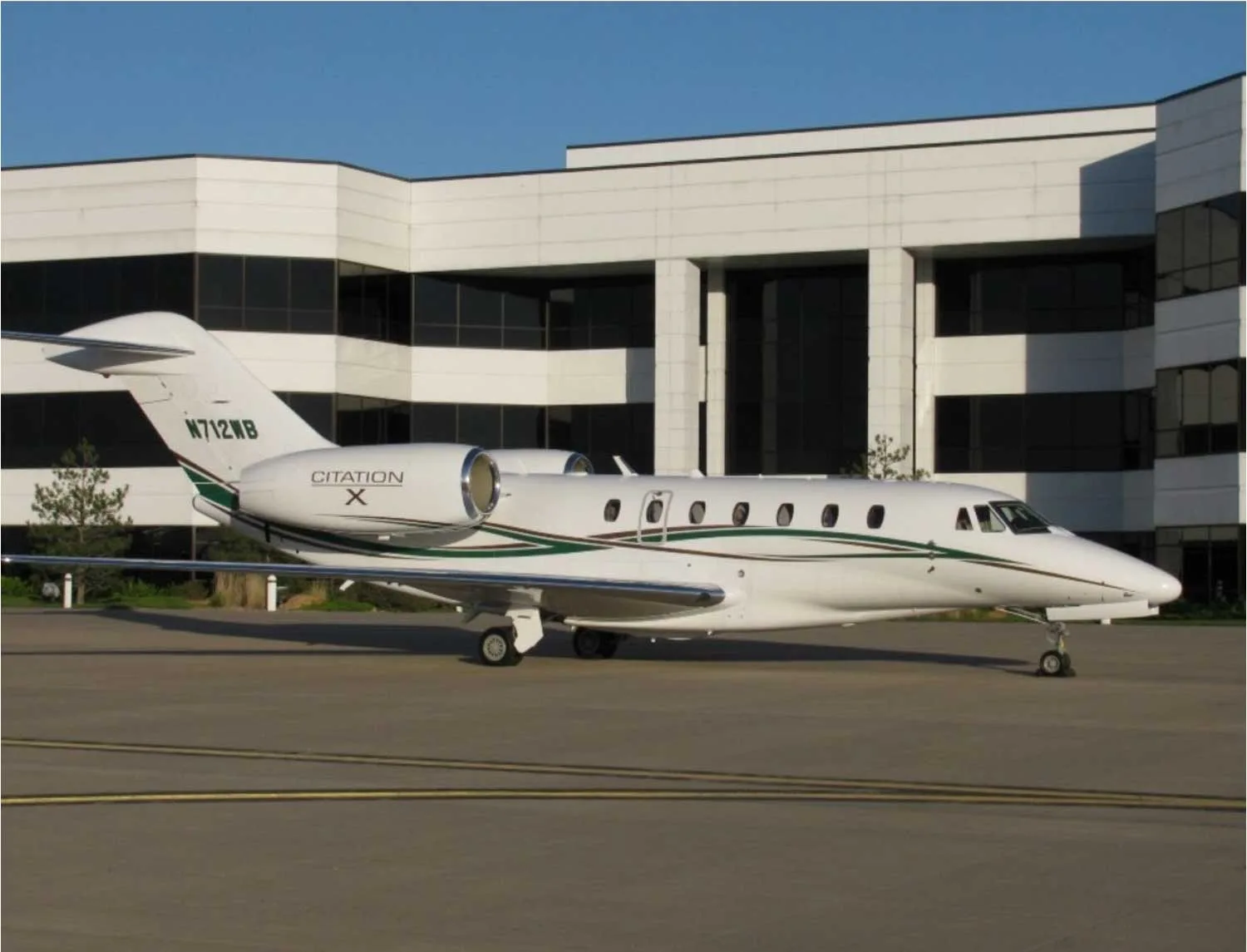 A white Cessna Citation X business jet with green and black stripes, registration N712WB, parked on an asphalt apron in front of a modern white building.