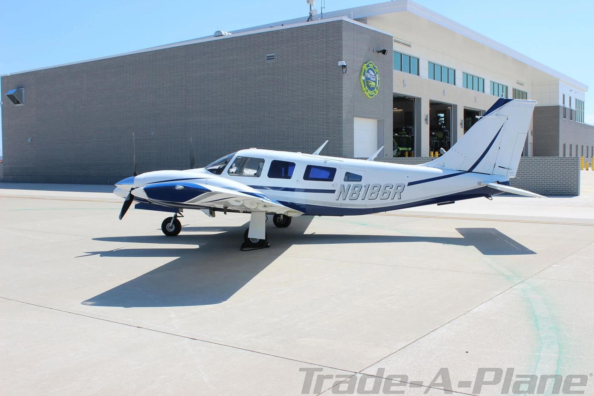 A side view of the white and blue Piper Seneca aircraft N8186R parked on a tarmac next to a grey building.