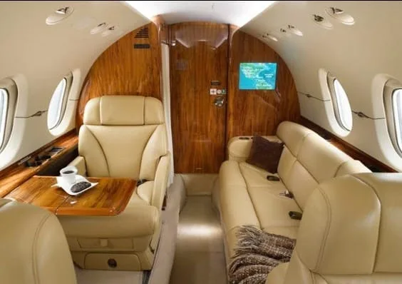 A view of the business jet cabin showing beige leather seats, a side-facing sofa, and a wooden partition.