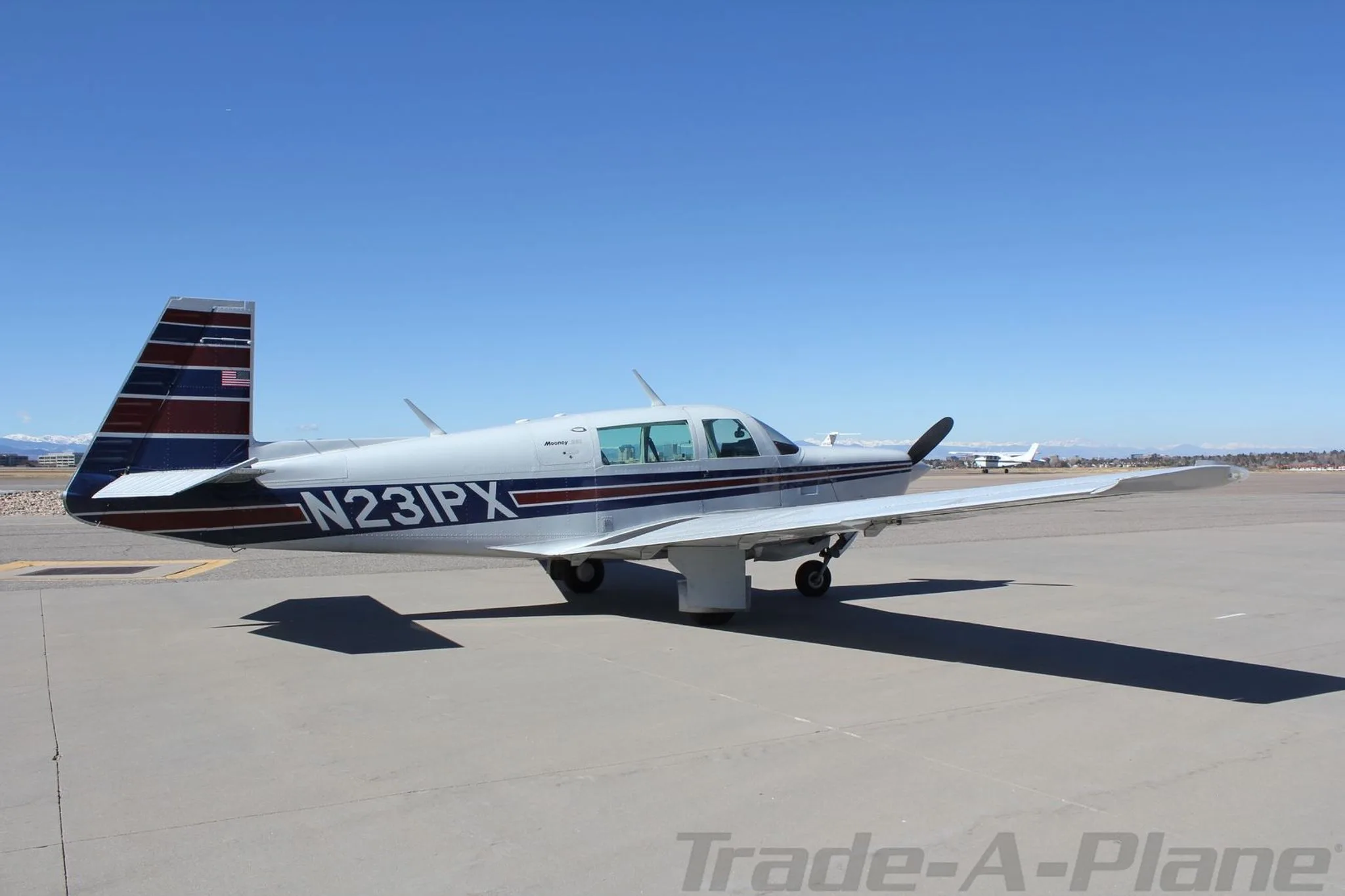 Side view of a silver single-engine aircraft with blue and red stripes, parked on an airfield with mountains in the distance.