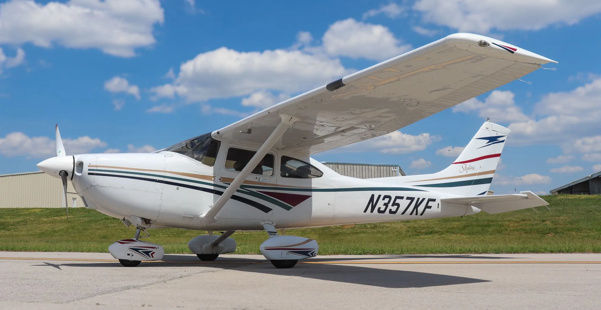 A white Cessna 182 Skylane with gold, green, and maroon stripes parked on an asphalt ramp.