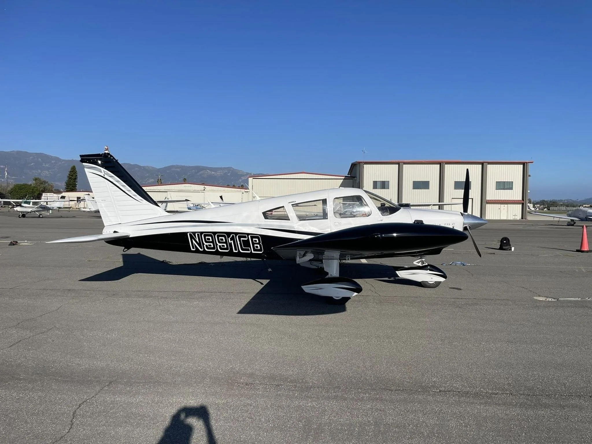 A side view of the white and black Piper Cherokee 235 C aircraft N991CB parked on an airfield next to hangars.