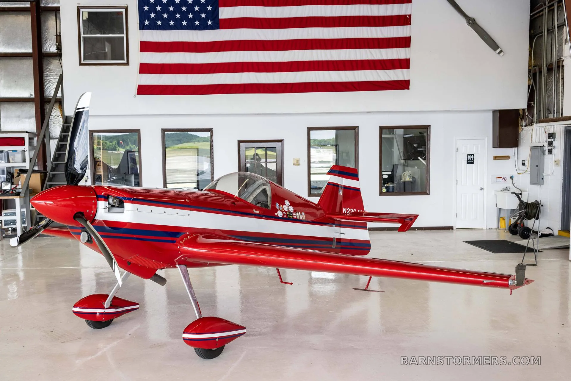 A red and white single-engine aerobatic aircraft with blue stripes, parked in a hangar under an American flag.