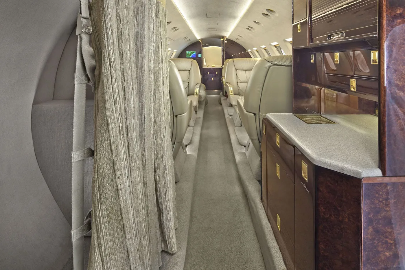 A view down the aisle of a private jet cabin, showing rows of cream leather seats and a dark wood galley area.