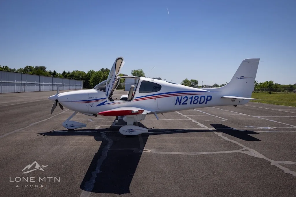 Side view of a white Cirrus aircraft (N218DP) with blue and red stripes and red wingtips, doors open, parked on an airfield.