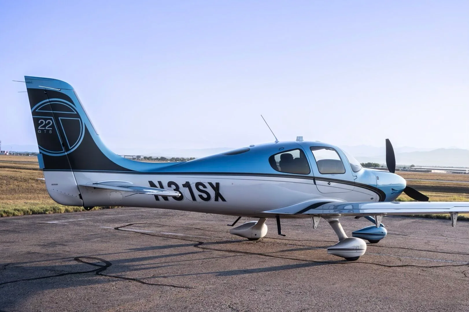 A Cirrus SR22T aircraft, N31SX, in blue and silver paint, parked on an airfield with distant mountains.