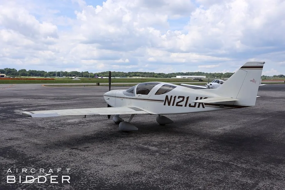 A white single-engine aircraft (N121JK) with brown stripes parked on an asphalt apron at an airfield.