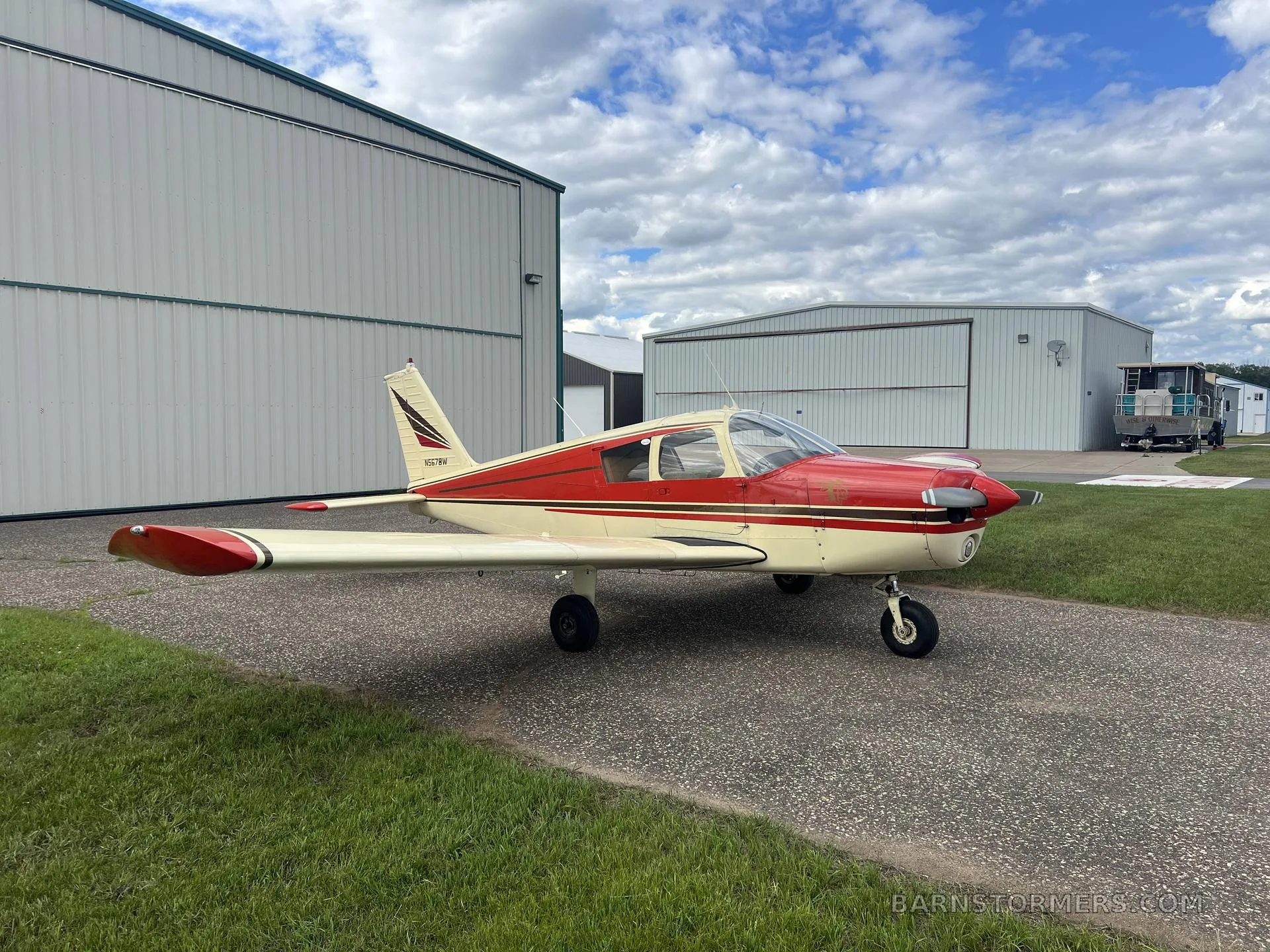 A red and cream single-engine propeller aircraft (N5678W) parked on an asphalt ramp next to hangars.