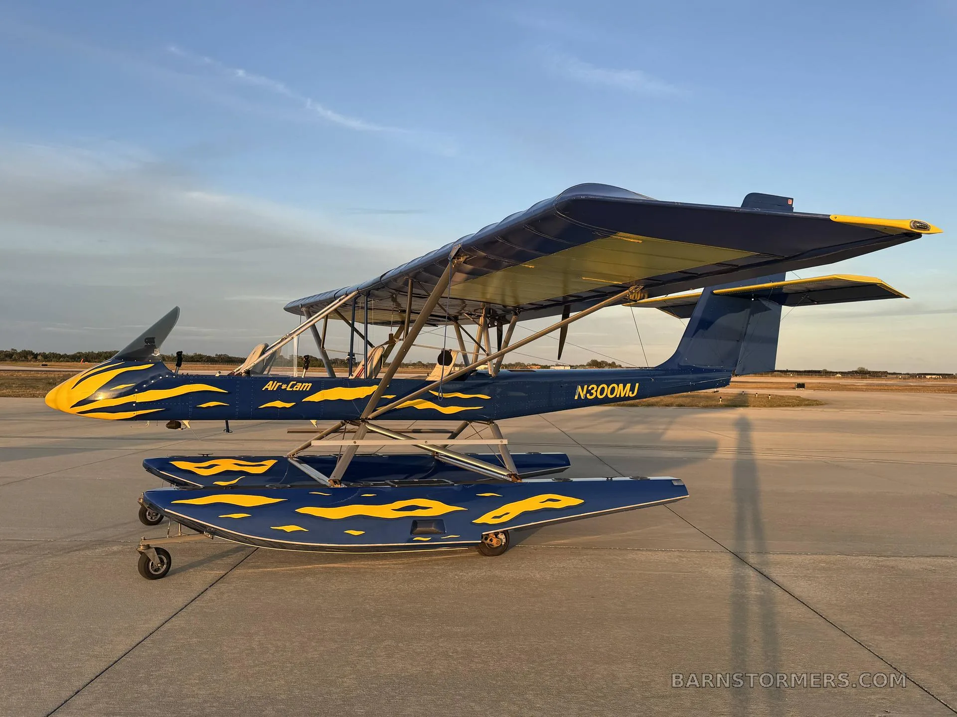 A side view of a blue and yellow Air-Cam seaplane with flame decals on floats, parked on an airfield at sunset.