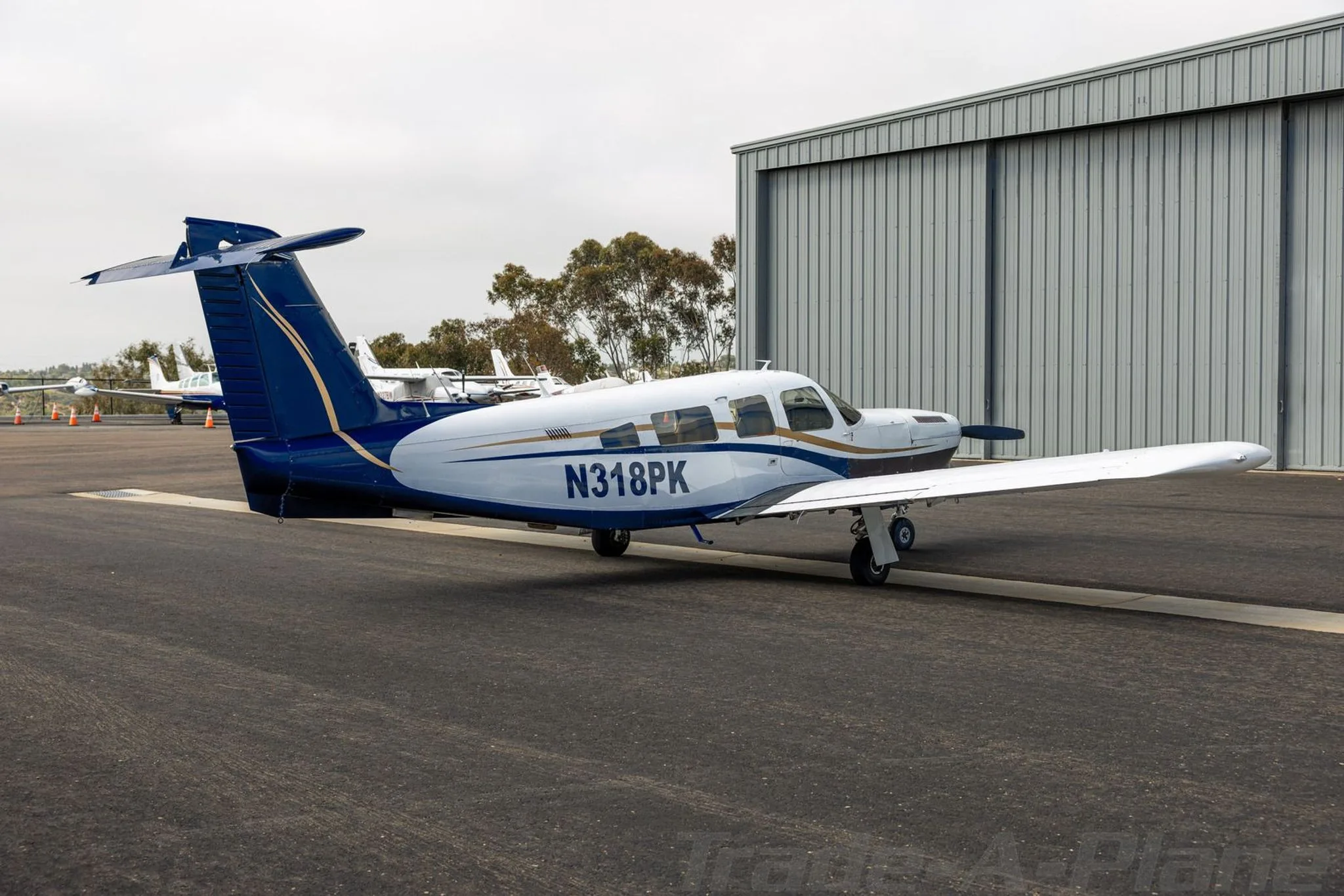 A three-quarter rear view of the white and blue Piper aircraft N318PK parked on an airport tarmac next to a hangar.