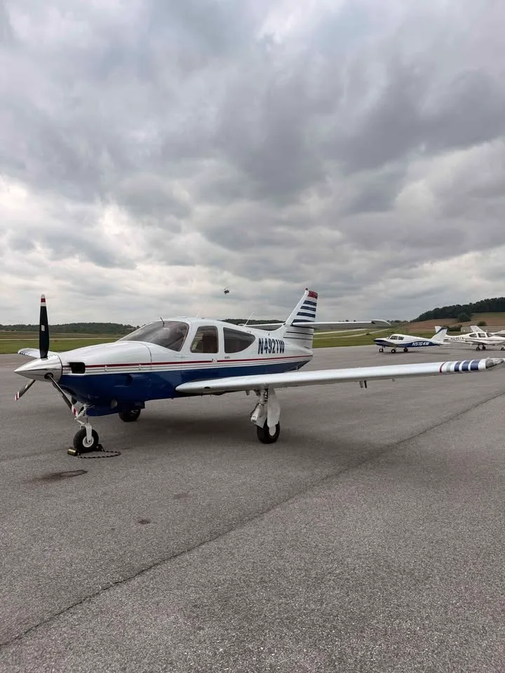 A side view of a white and blue single-engine aircraft with registration N4921W parked on the tarmac with other aircraft in the background.