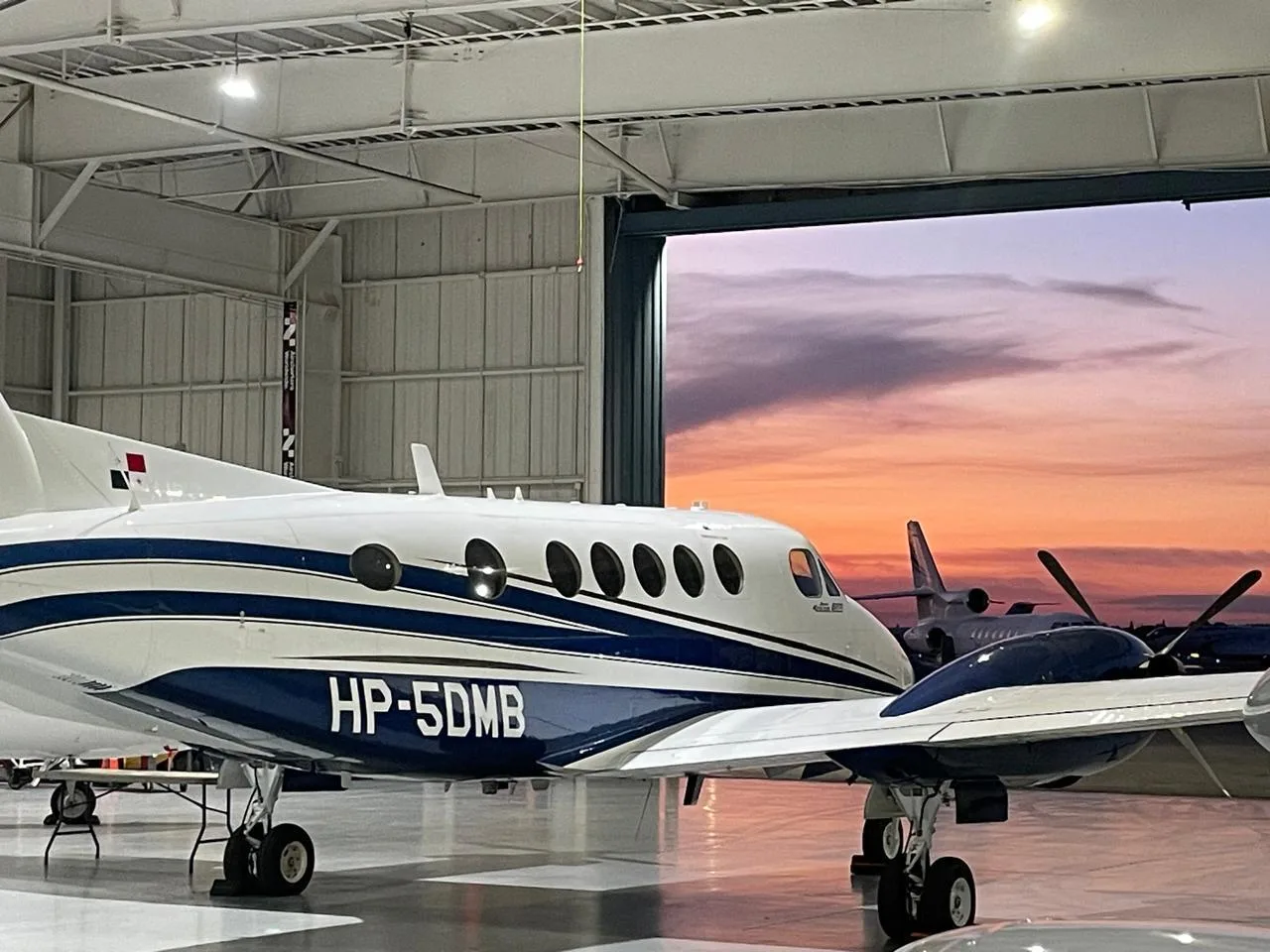 A white and blue turboprop aircraft (King Air) with registration HP-5DMB parked inside a hangar, with a colorful sunset visible through the open hangar door.