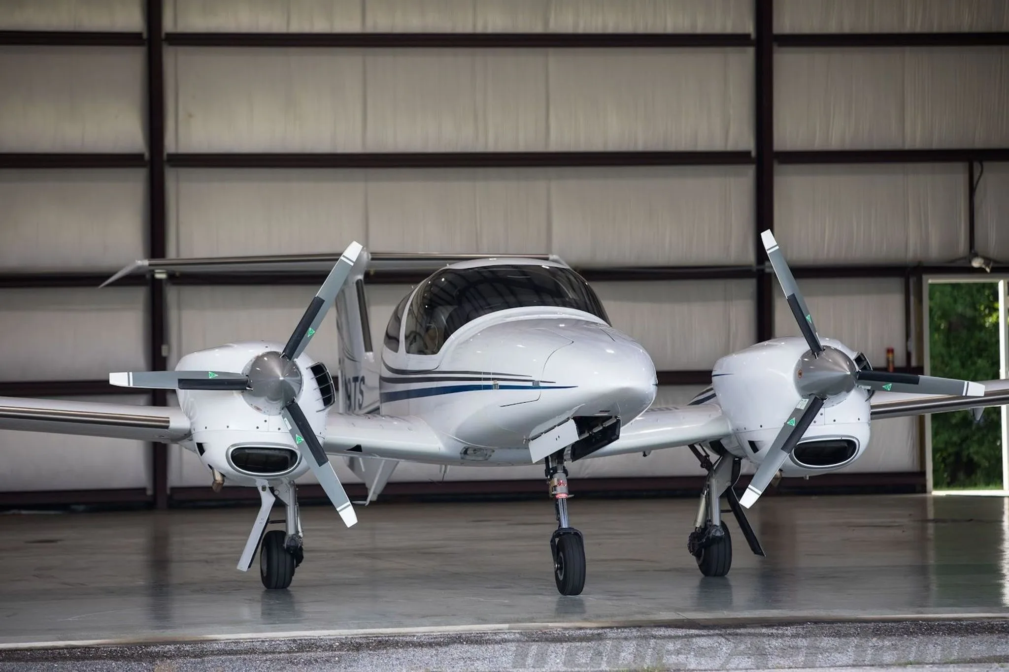 A white Diamond DA42-NG aircraft with blue and grey stripes is parked inside a hangar, viewed from the front.