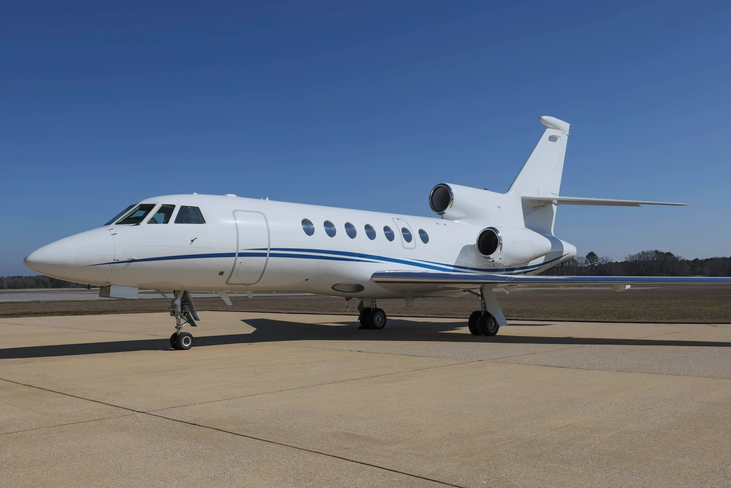 A white Dassault Falcon 900EX business jet with blue stripes is parked on a tarmac under a clear blue sky.