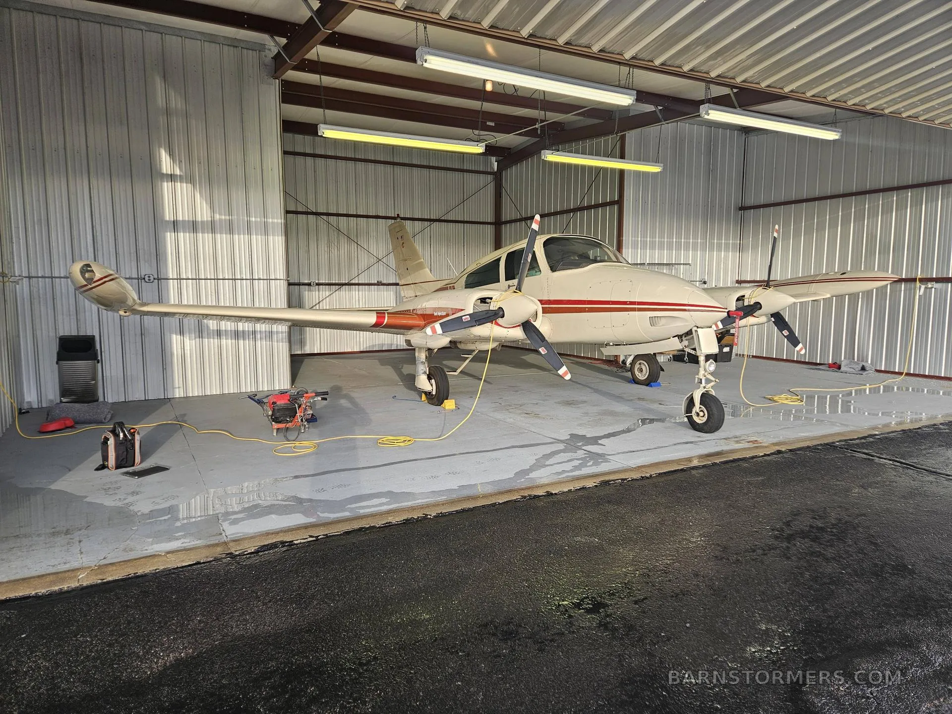 A white and red twin-engine propeller aircraft is parked inside a hangar with its propellers tied down.