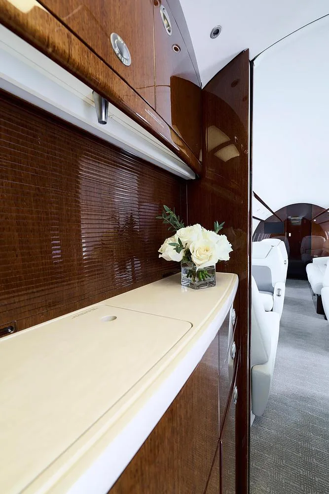 A close-up view of a polished dark wood cabinet with a light-colored countertop and a vase of white flowers in a business jet cabin.