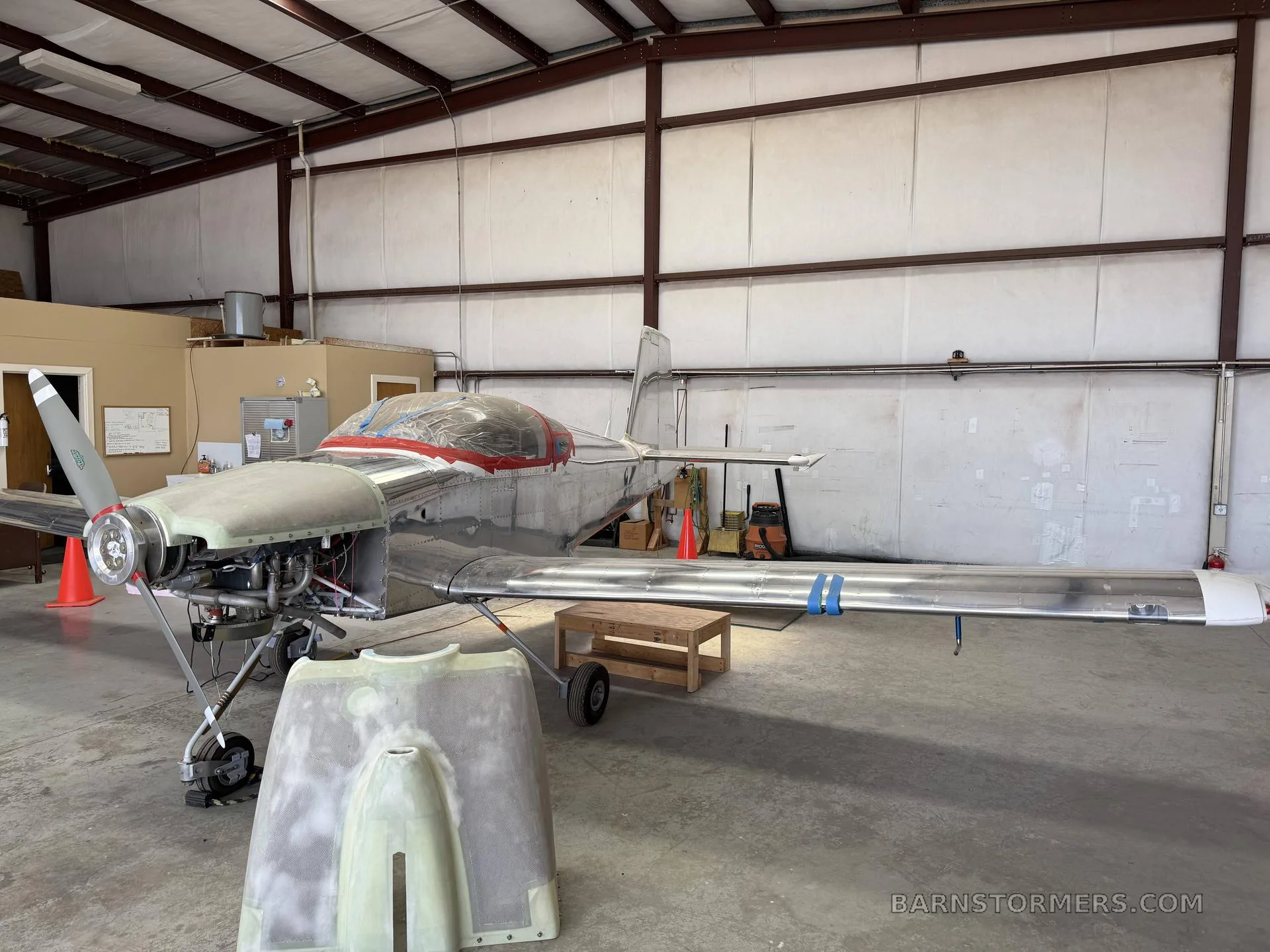 A partially assembled, unpainted metal aircraft with a red-taped canopy and exposed engine in a hangar.