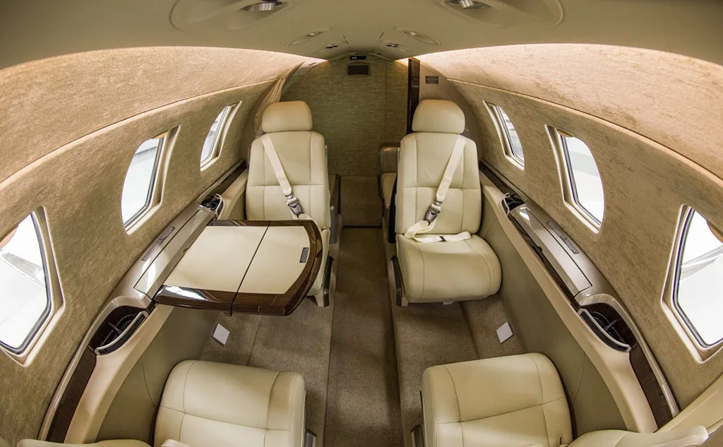 The beige interior cabin of a private jet, showing four passenger seats and a fold-out table.