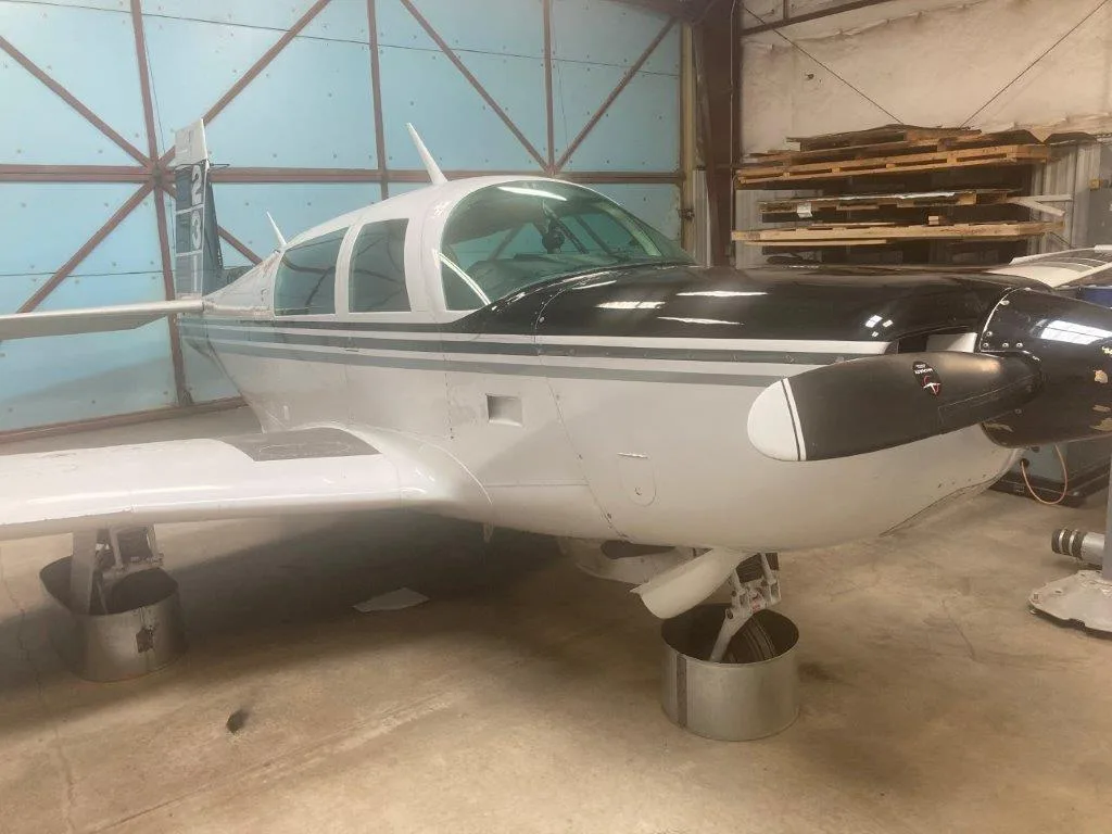 Front-side view of a white and black Grumman American AA-5 Traveler aircraft, N1047A, on stands inside a hangar.