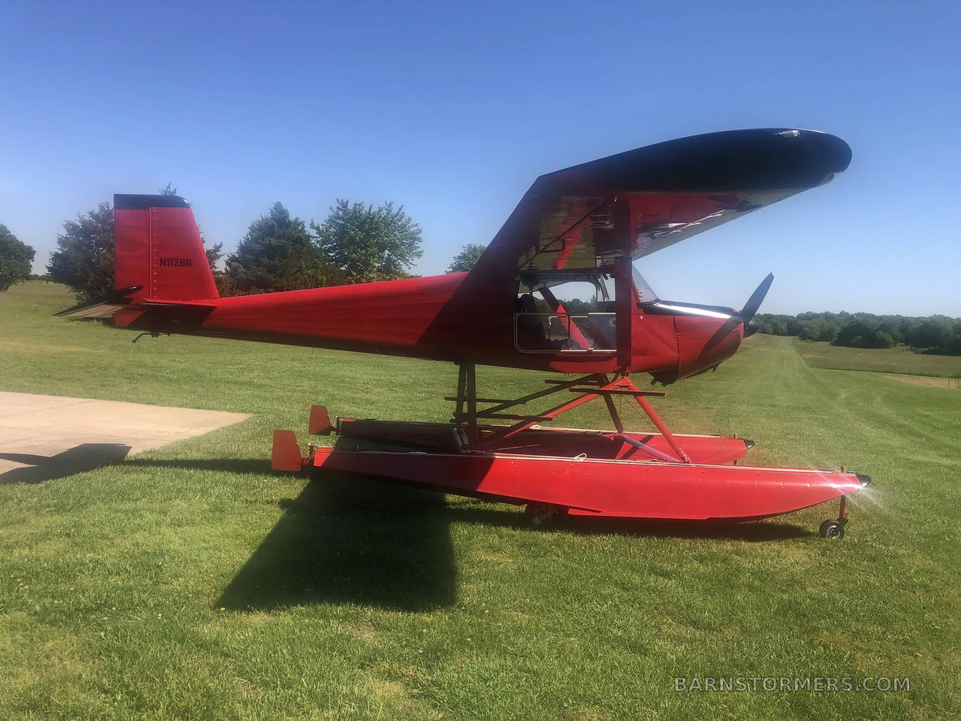 A red floatplane with registration N11286 is parked on green grass next to a concrete pad under a clear blue sky.