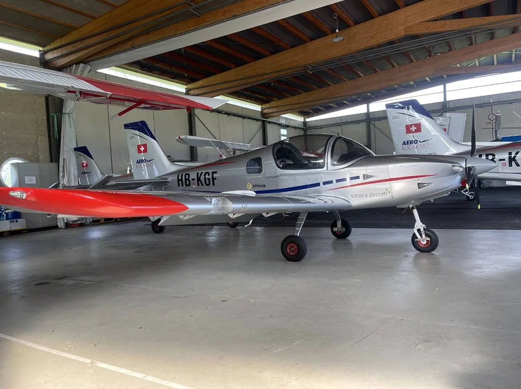 A silver Sonaca aircraft with red wingtips and registration HB-KGF parked in a hangar.