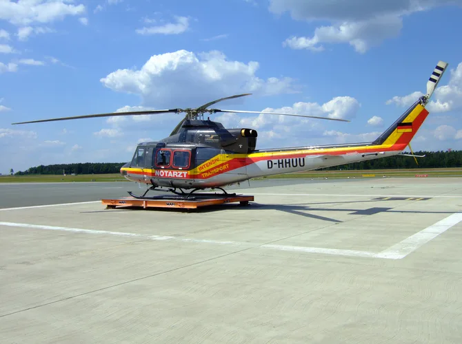 A black and silver air ambulance helicopter with red and yellow stripes and German flag colors on the tail, parked on a concrete apron under a blue sky.