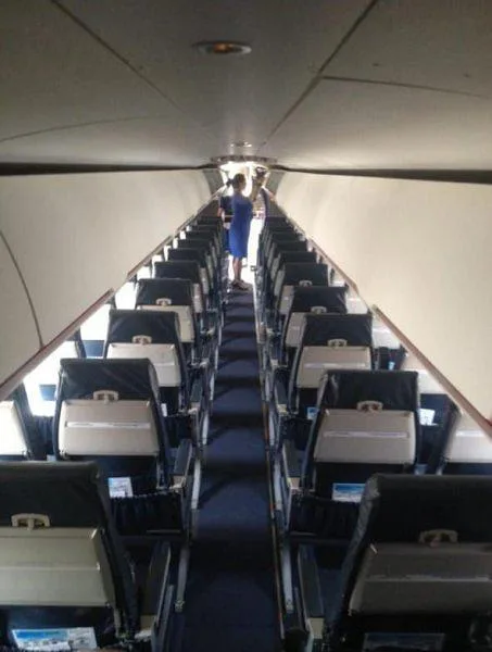 The interior cabin of an aircraft with rows of dark seats and a person in a blue uniform standing at the rear.