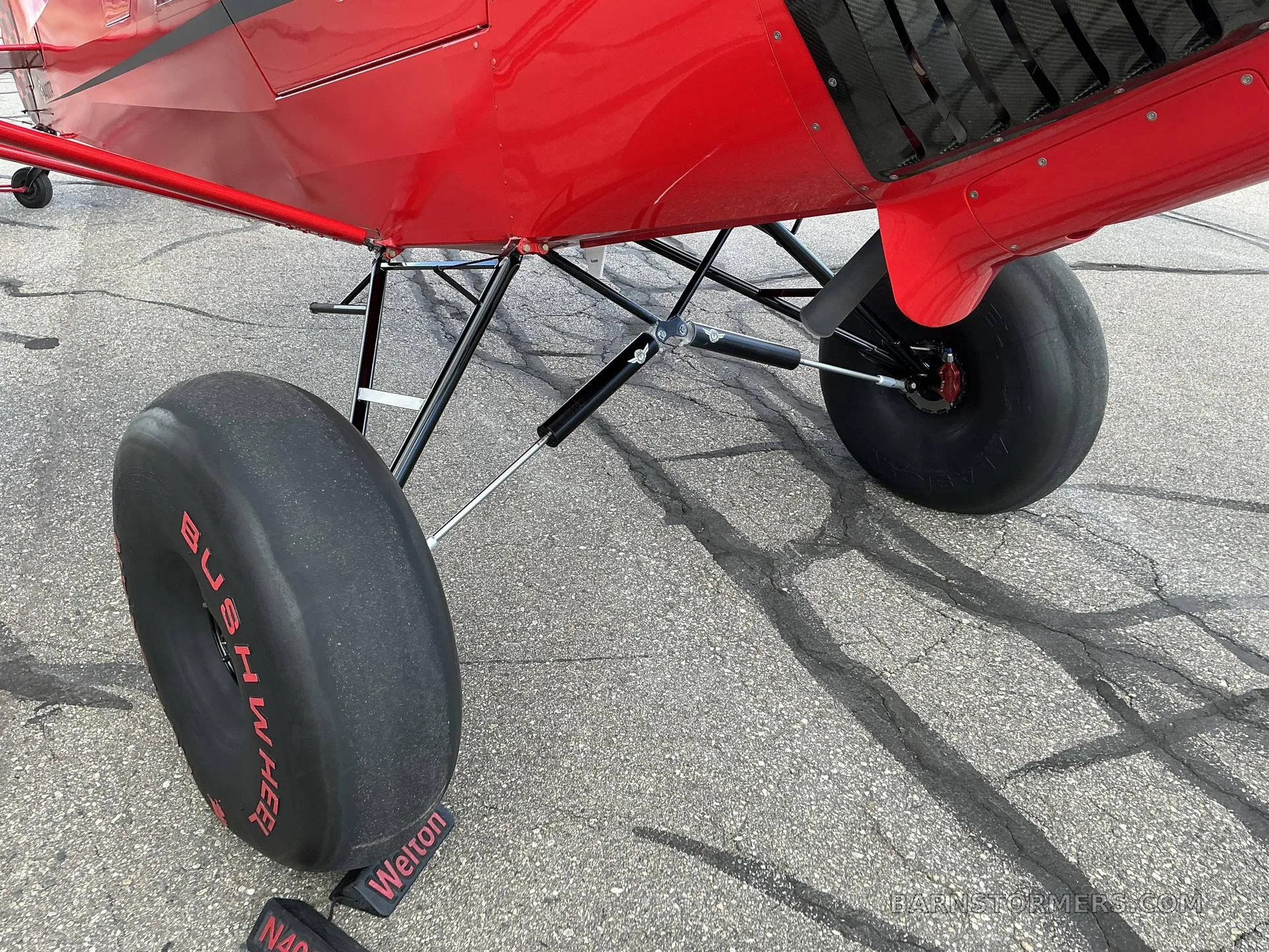 A close-up view of the robust landing gear and large "Bushwheel" tires of a red aircraft on an asphalt surface.