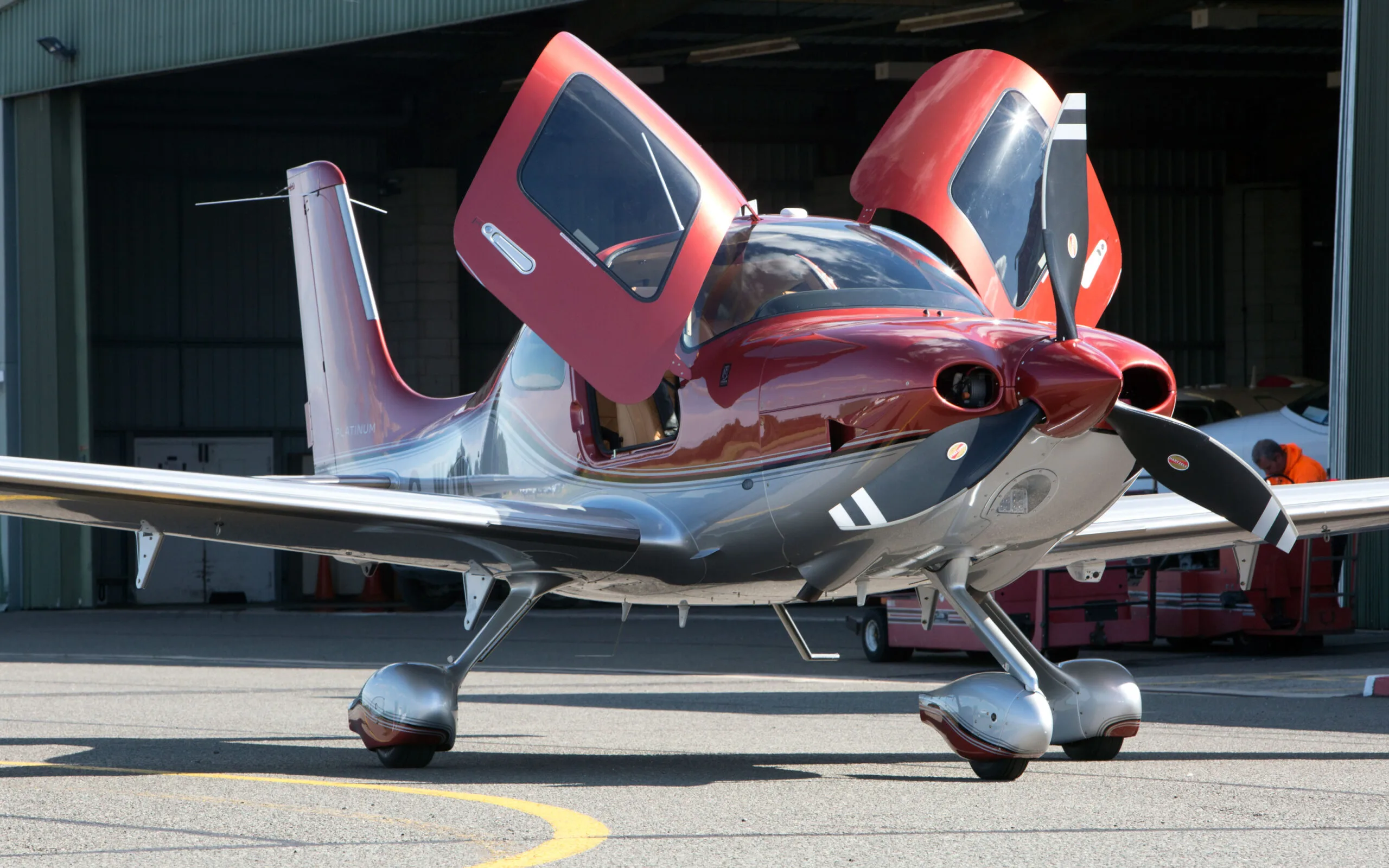 A red and silver Cirrus SR22T GTS aircraft with its gull-wing doors open, parked on an airfield tarmac in front of a hangar.