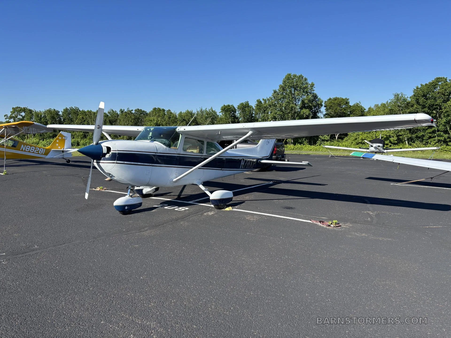 A white and blue Cessna 172 (N77XP) with a propeller, parked on an airfield next to a yellow aircraft.