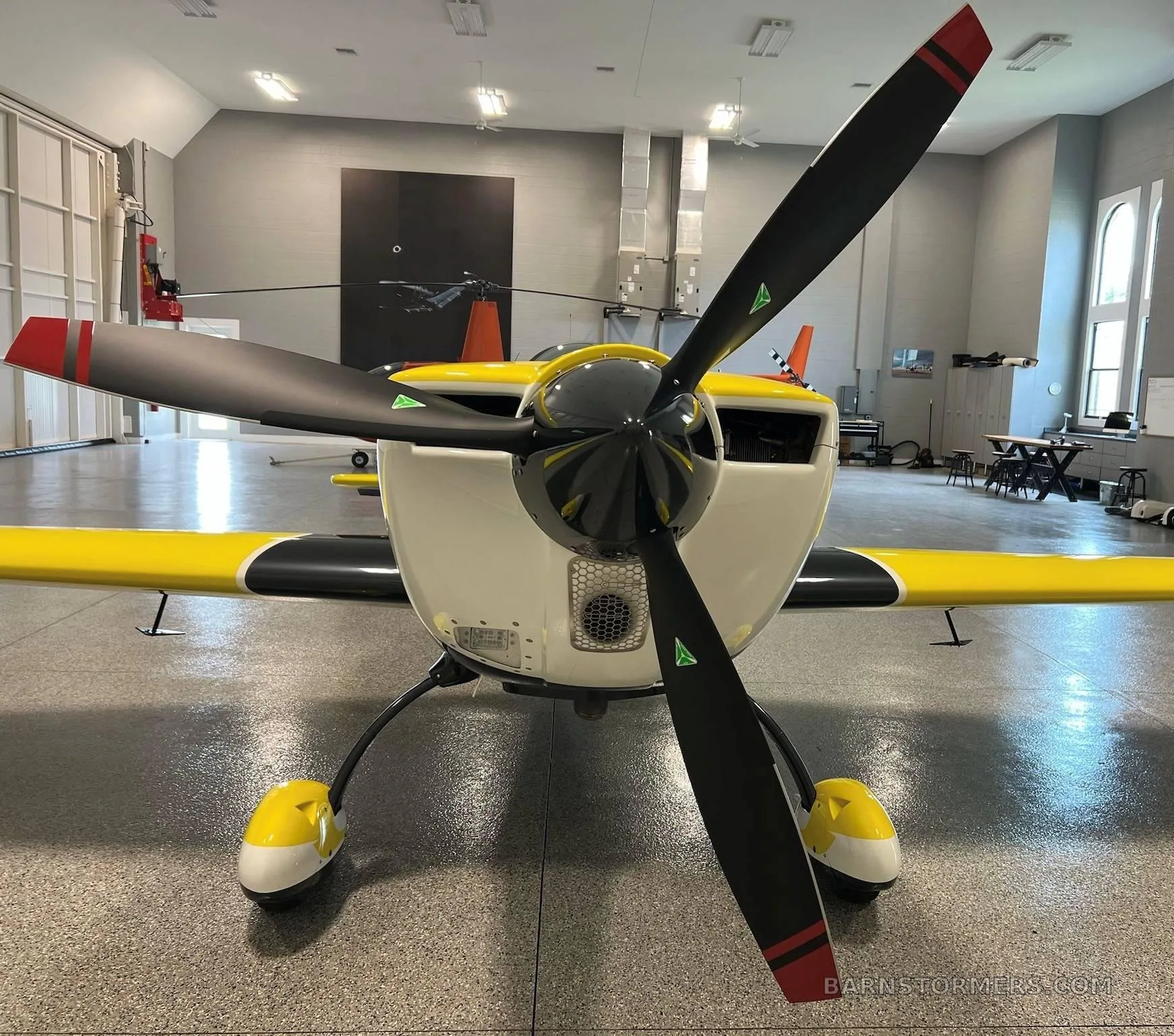 The front view of a yellow and white Extra 330LT aerobatic aircraft with a three-bladed propeller, in a hangar.