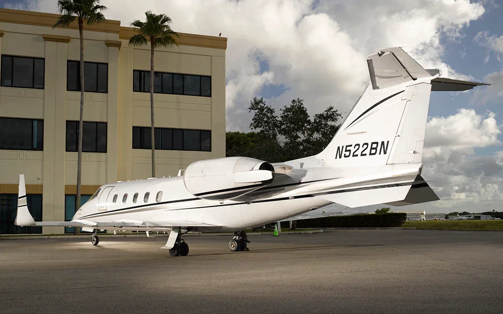 A white Learjet with black and grey stripes, tail number N522BN, parked next to a tan building with palm trees.
