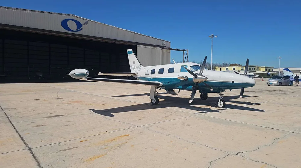 A Piper PA-31 Navajo, registration N444PC, in a white and teal paint scheme, parked on a tarmac with a hangar in the background.