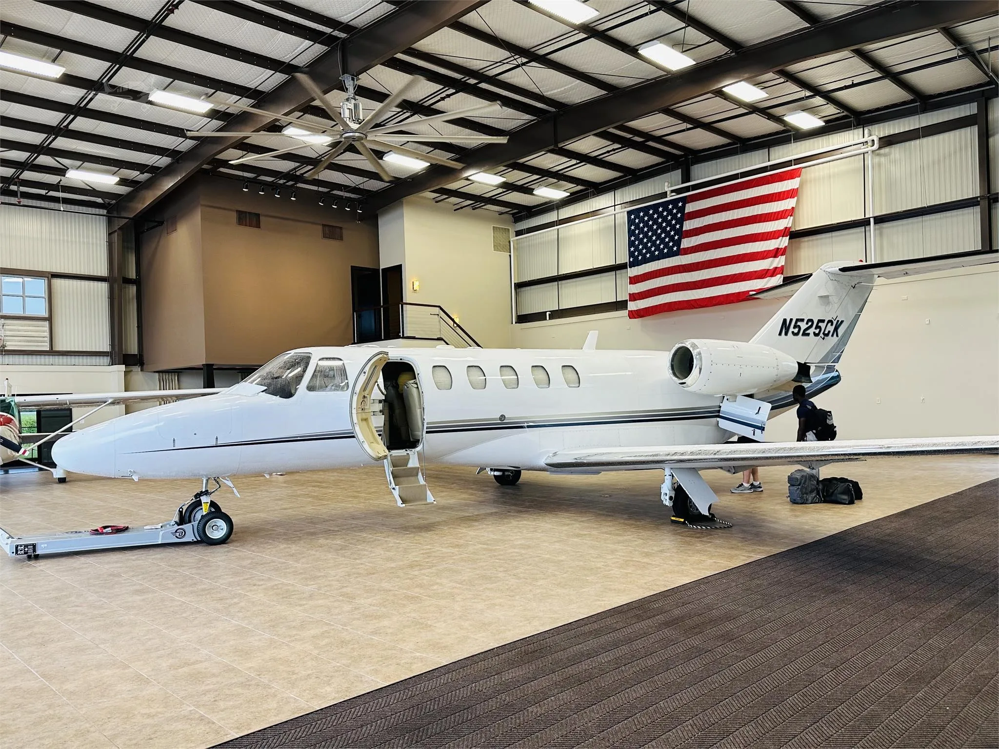 A white Cessna CitationJet with tail number N525CK is parked in a hangar with its door open, an American flag hangs in the background.