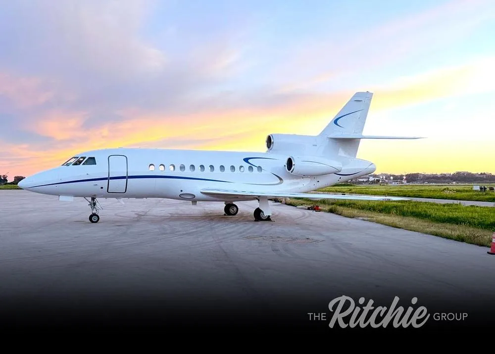 A white Dassault Falcon business jet with blue accents parked on a tarmac at sunset.