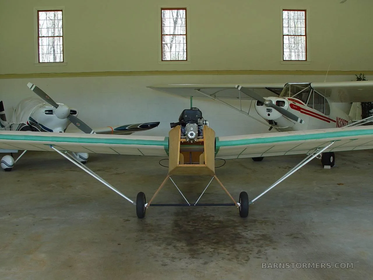 A front view of a light aircraft with a wooden fuselage and green trim, parked in a hangar next to another white aircraft.