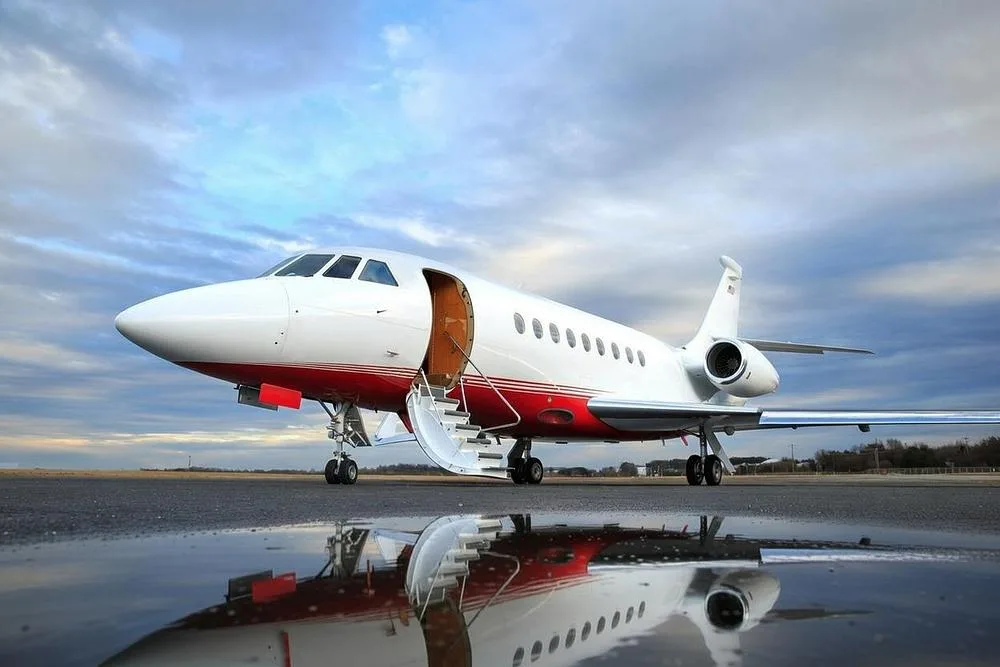 A white and red Dassault Falcon 900 private jet with its entry door open and stairs extended, reflecting in a puddle on the tarmac.