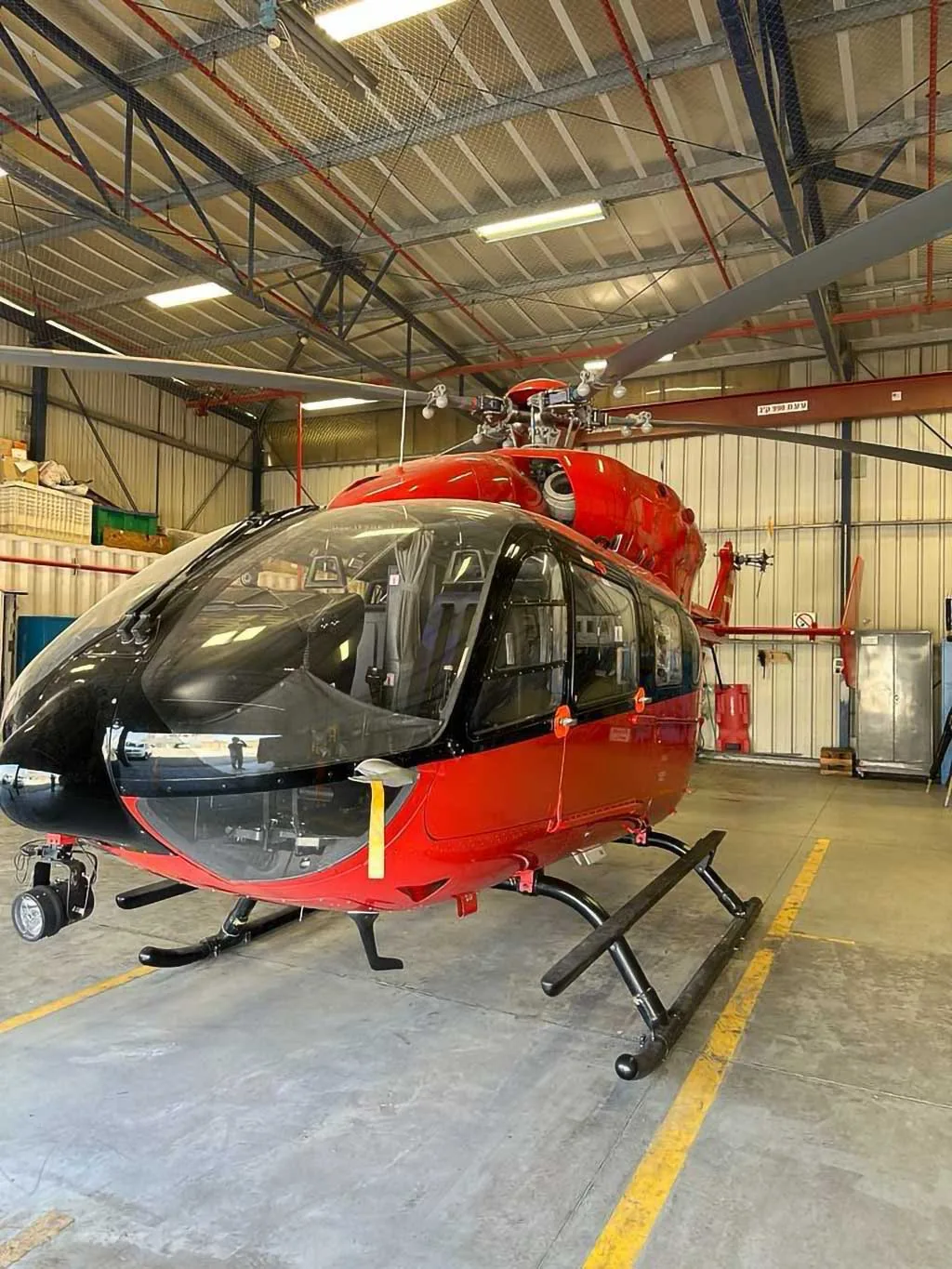 A red Airbus H135 helicopter with black skids and a camera mounted on the nose, parked inside a hangar.
