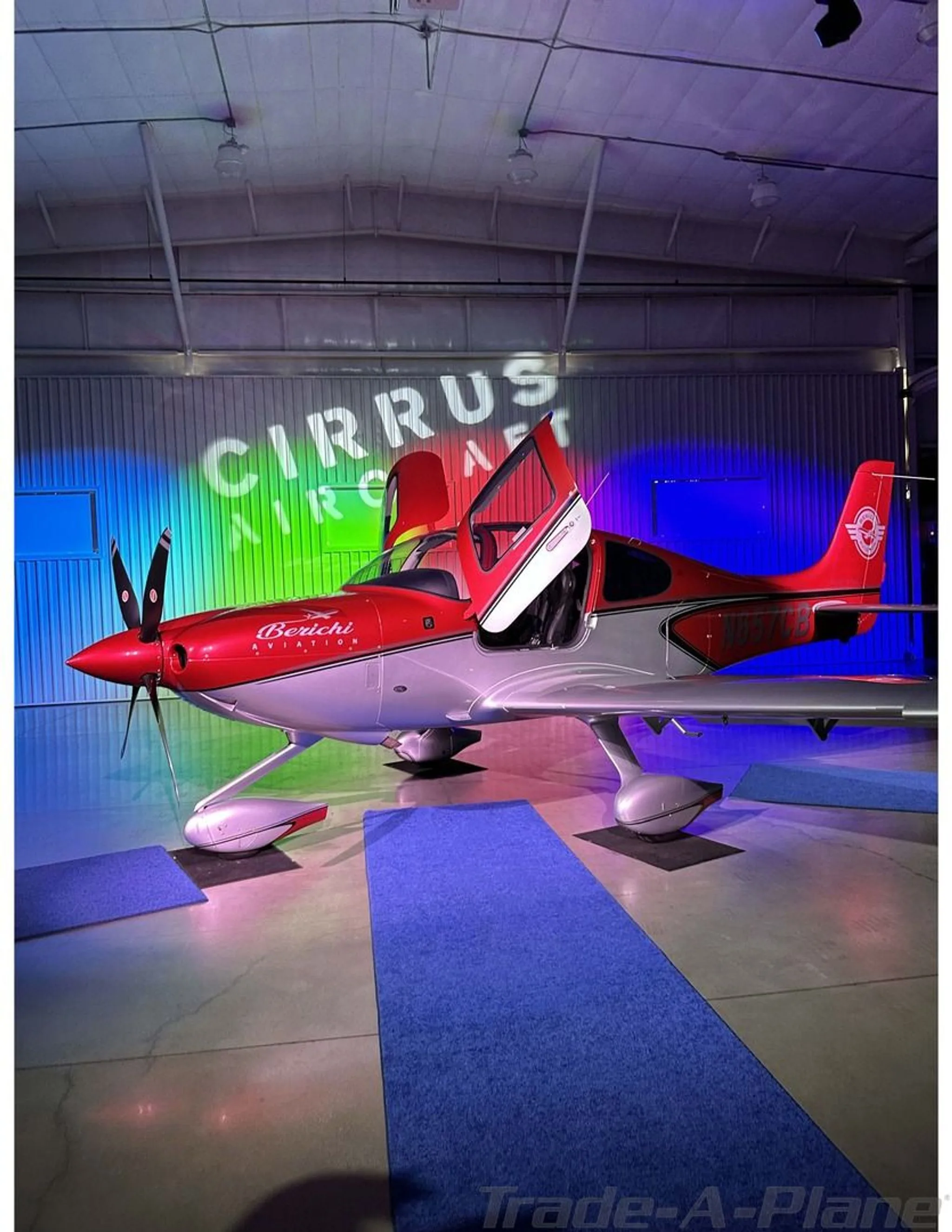 A red and silver Cirrus SR22 aircraft, N657CB, with its doors open, displayed in a hangar with "Cirrus Aircraft" projected on the wall.