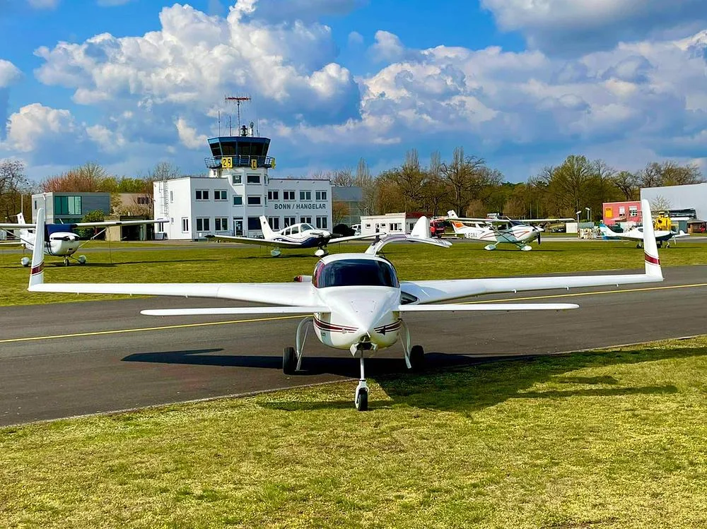 A white Velocity Elite aircraft with red and gold stripes parked on an airfield in front of a control tower.